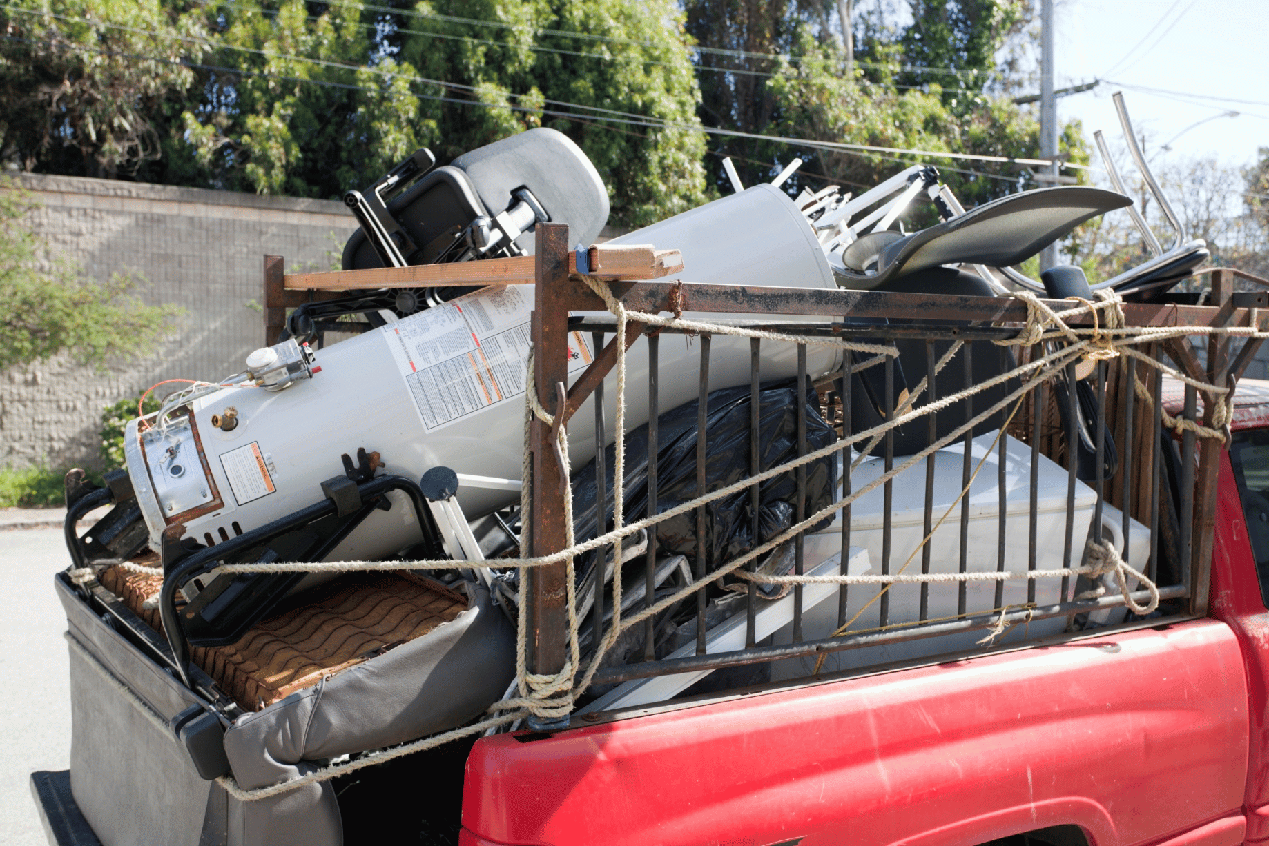 Red pickup truck bed overflowing with junk—furniture, appliances, and a water heater strapped down with thick rope