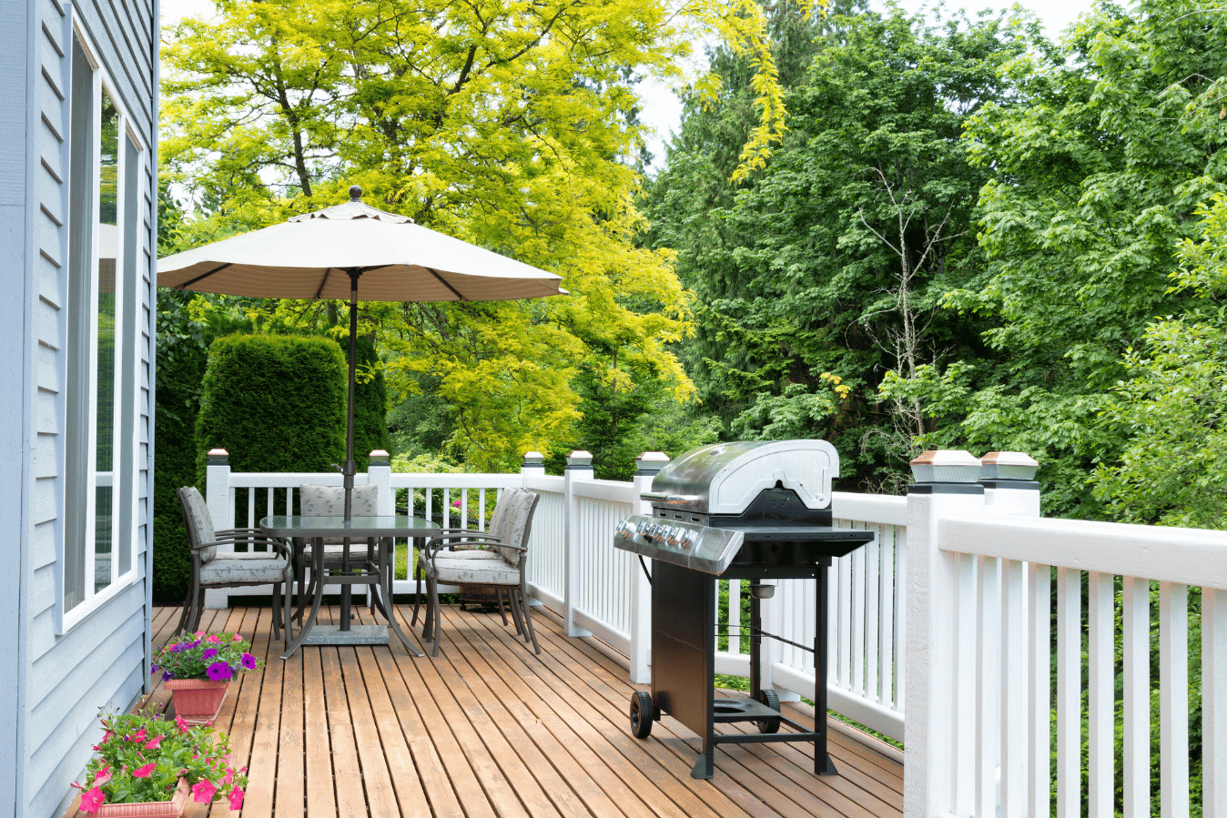 Wood deck with a round dining table, four cushioned chairs, and a large beige umbrella, beside a stainless gas grill and white railing, green trees beyond.